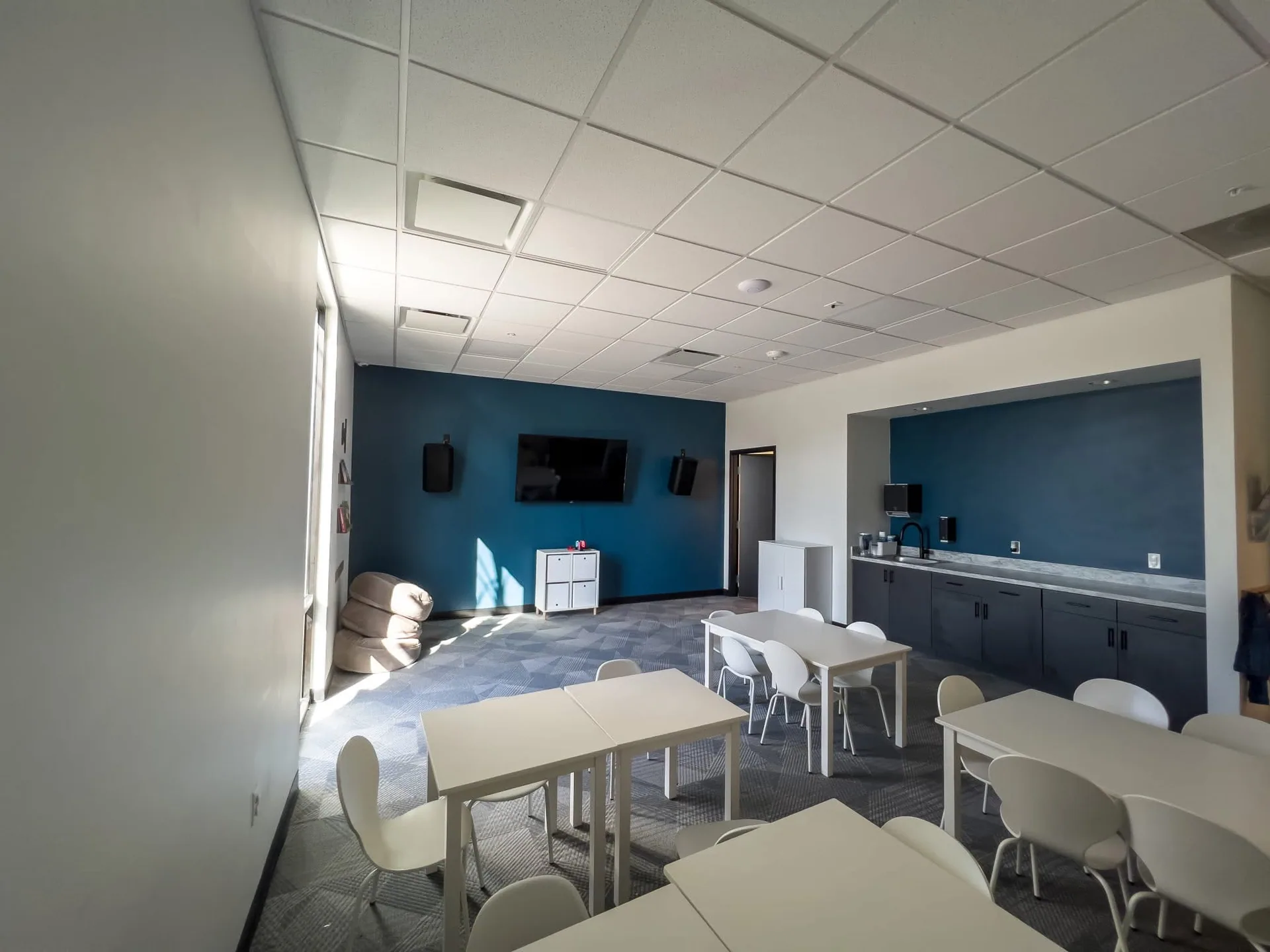 Classroom with a blue accent wall, tables and chairs, and a ceiling grid with acoustical tiles.